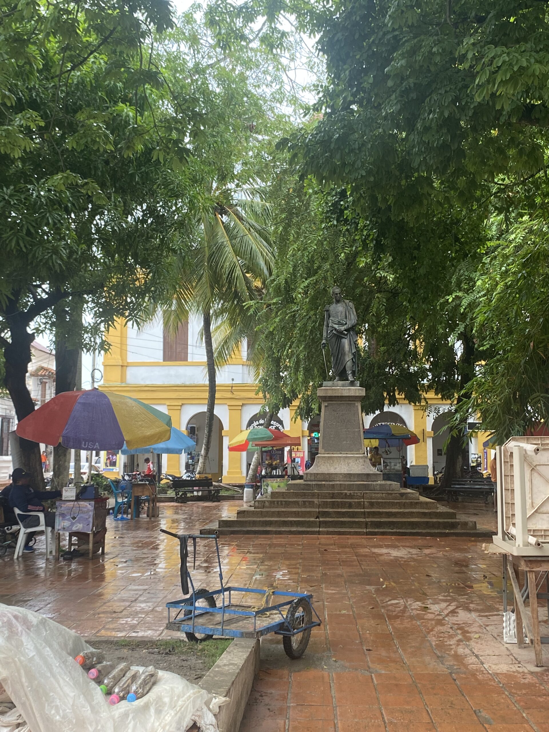 Plaça de Mompox amb arbres, para-sols de colors i una façana groga al fons