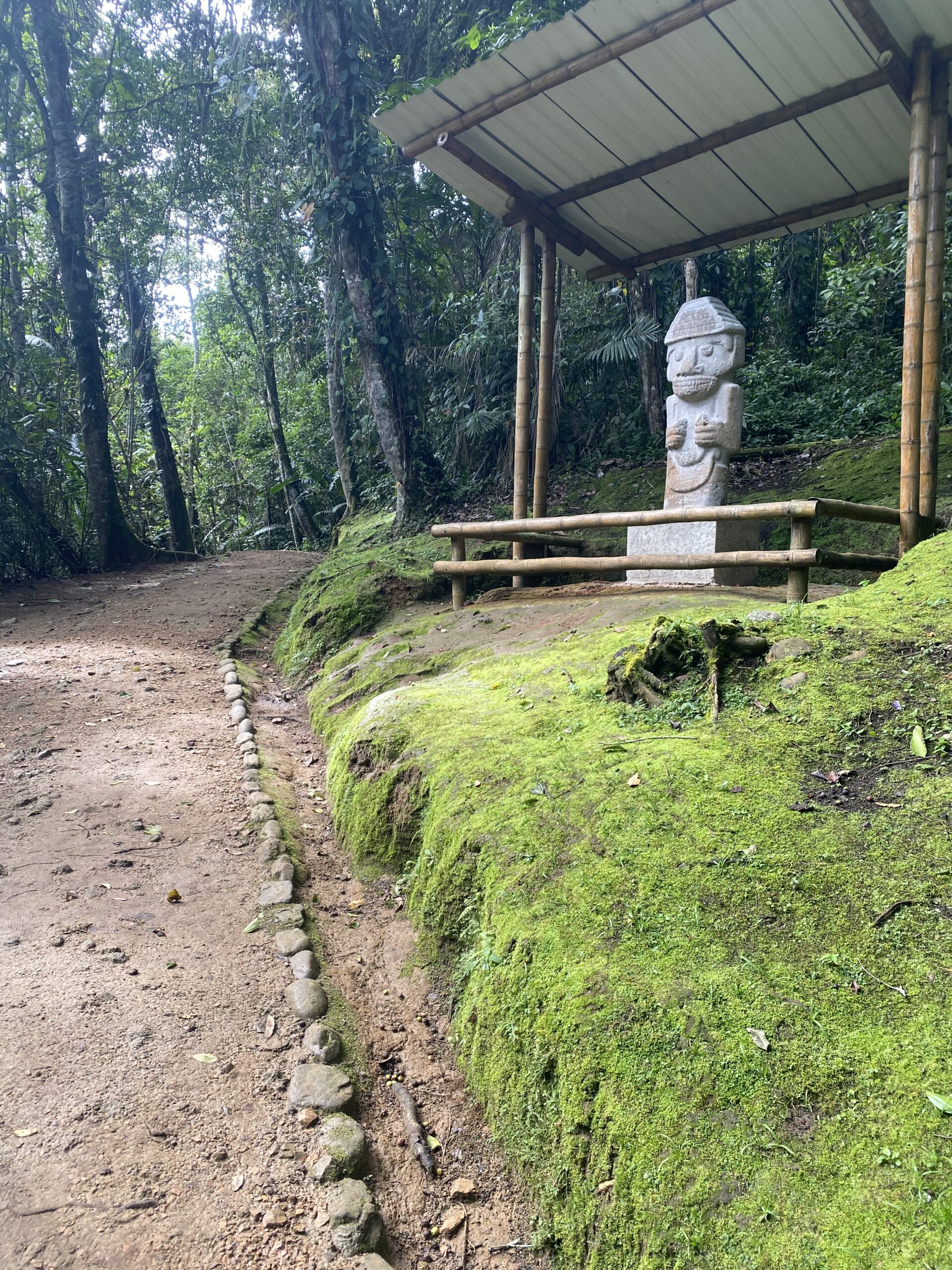 Estàtua de pedra precolombina damunt d'un túmul d'herba, sota un cobert i envoltada d'arbres alts